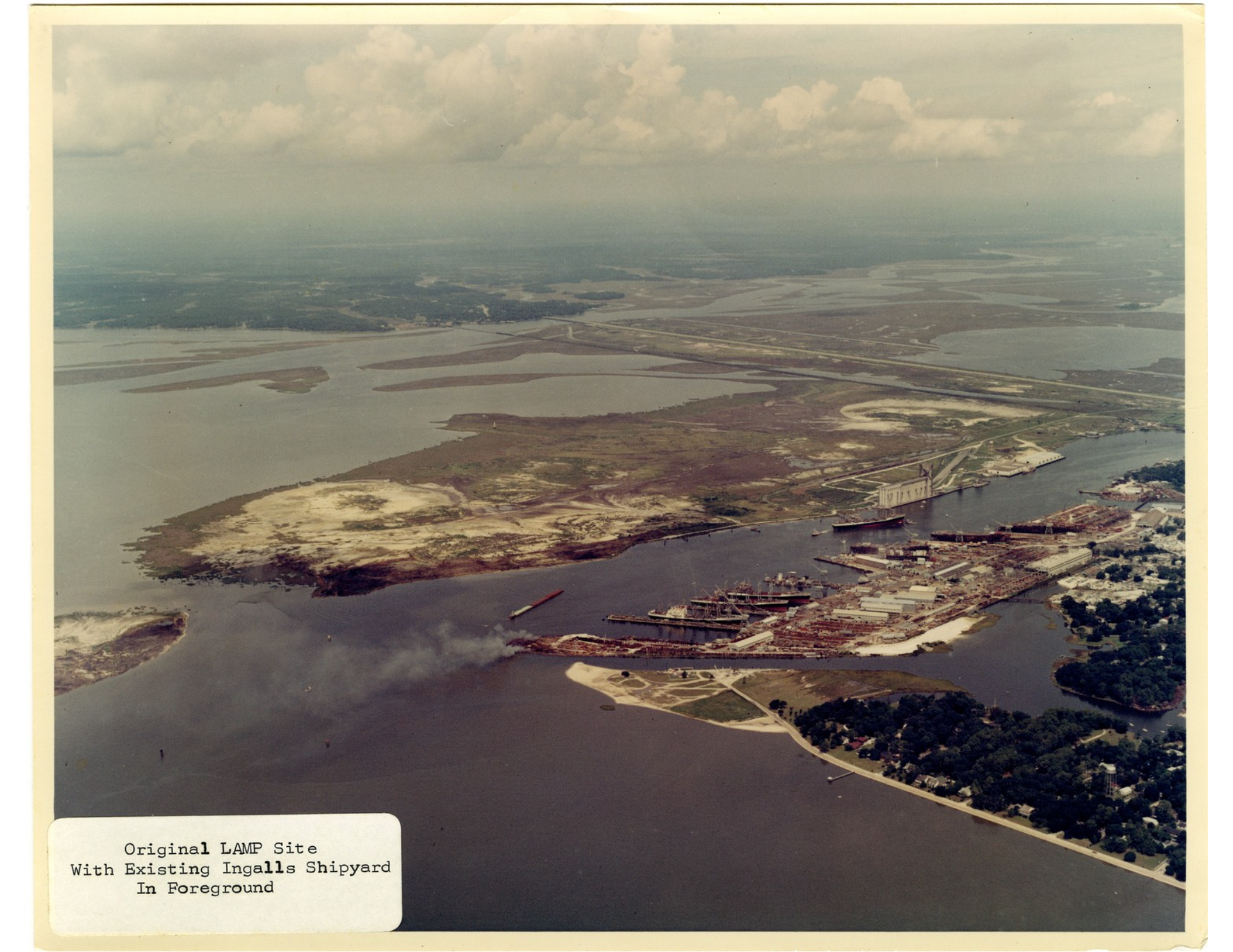 Color aerial photograph of coastal industrial area with waterways, islands and shipyard facilities. A small caption in the lower left reads, "Original LAMP Site With Existing Ingalls Shipyard In Foreground."