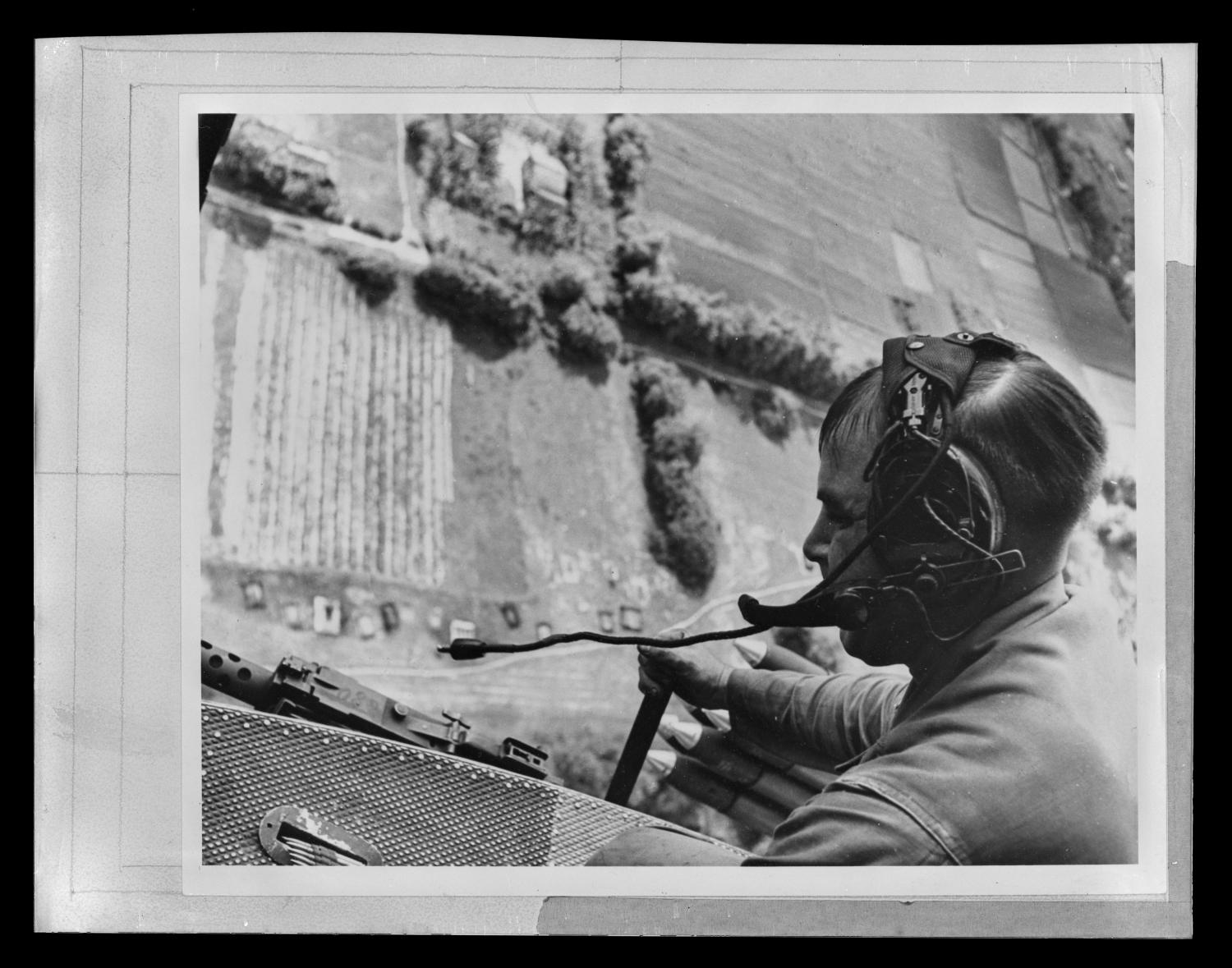 Photograph of a door gunner leaning out of a UH-1A Iroquois helicopter. He wears a headset and wears a long-sleeved, collared shirt. The pastures, roads, and trees can be seen on the ground far below, 1970s.